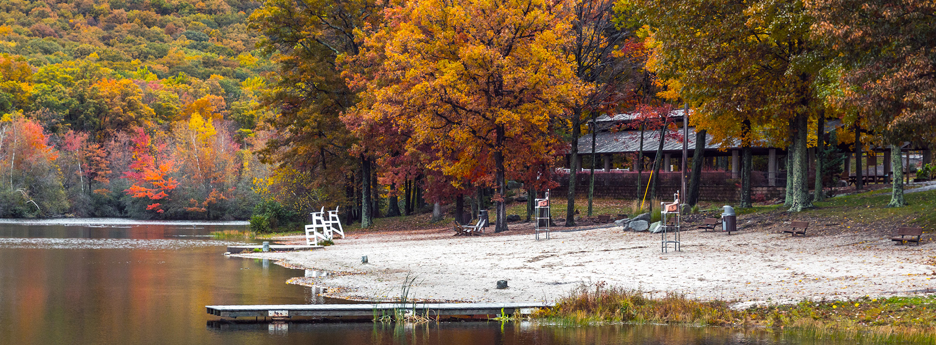 The image is a panoramic view of a serene autumn landscape featuring a lake, trees with fall foliage, a dock, and a beach area.