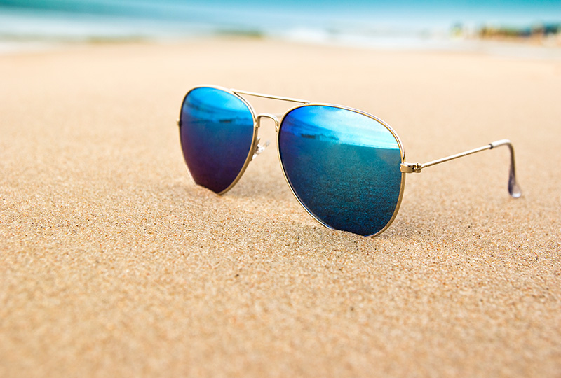 A pair of sunglasses with blue lenses resting on a sandy beach.
