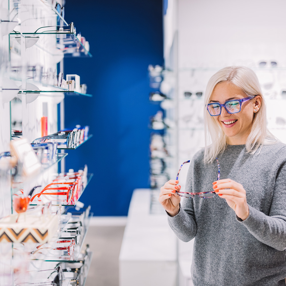 A woman with glasses and a blue sweater is standing in front of an optical shop display, holding a pair of glasses.