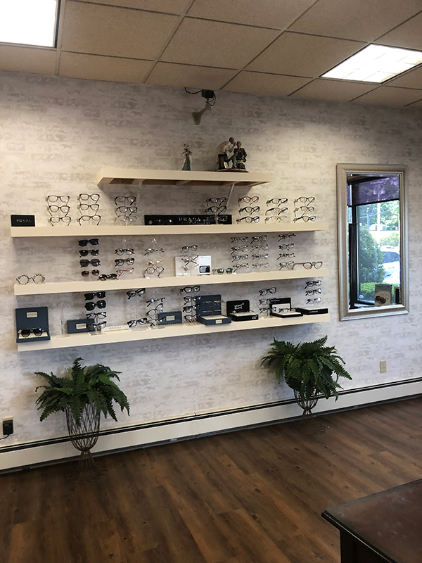 An optical shop with a large display of eyeglasses on shelves, set against a white brick wall and a window with blinds.