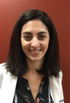 The image is a photograph of a woman standing in front of a red wall, smiling and posing for the camera. She has short dark hair, wears glasses, and is dressed in professional attire including a white blazer over a black top and a patterned skirt.