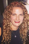 The image shows a woman with long curly hair, wearing a black top and a necklace, smiling at the camera.