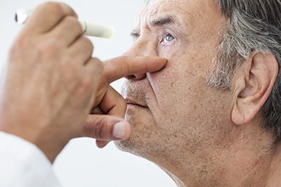 The image shows a man receiving an eye exam, with his nose gently placed on the tip of a device held by a professional.