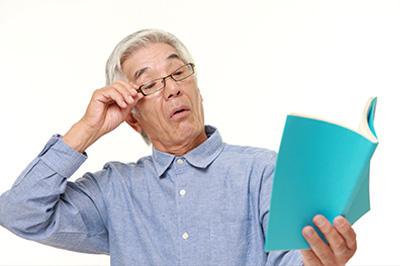 An elderly man in a blue shirt, wearing glasses and holding an open book with both hands, appears to be reading or studying.