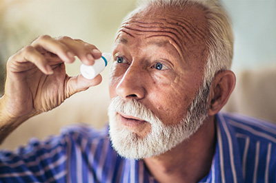 The image depicts an elderly man with a white beard and mustache, wearing glasses, looking intently at a magnifying glass through which he is examining something. He appears to be in a contemplative or focused state while observing the object in the glass.