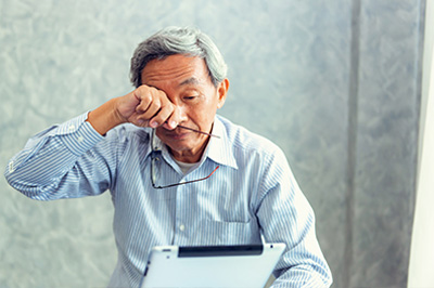 An older man in a blue shirt, seated at a desk and using a laptop with his eyes closed, appearing to be engaged in work or a personal task.