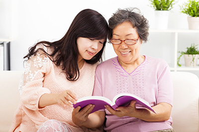 A woman and an older woman are sitting on a sofa, both engrossed in reading a book.