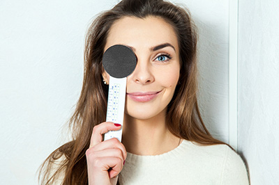 A woman holding a thermometer to her forehead, with a magnifying glass placed over her eye.